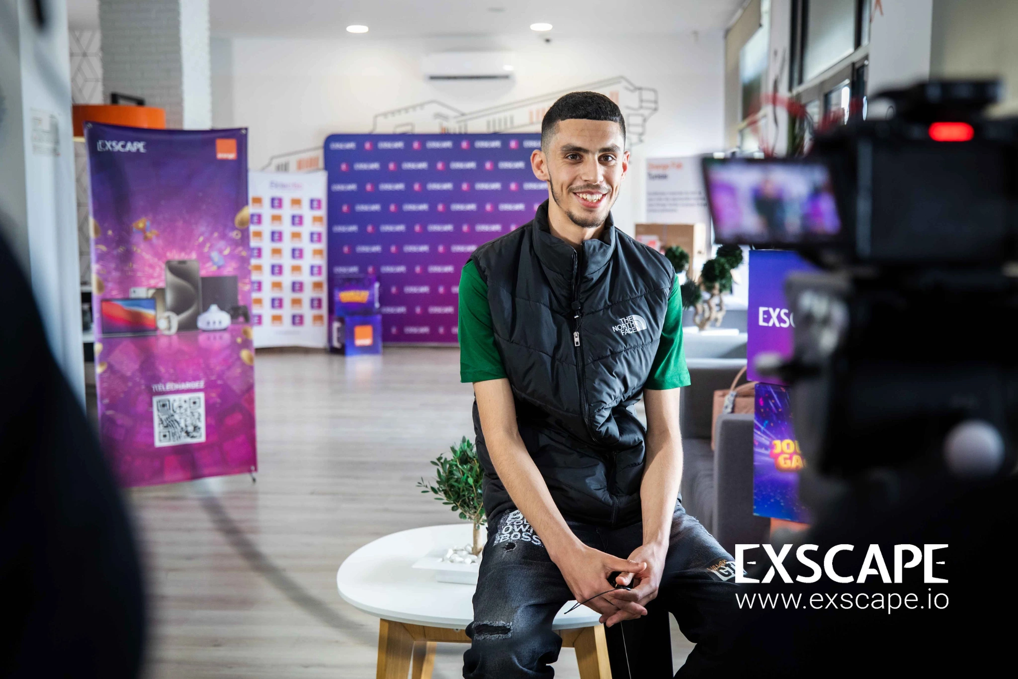 A top winner sits on a stool and looks into the camera feeling proud as the Orange prize venue is in the background. a camera is on a tripod in the foreground